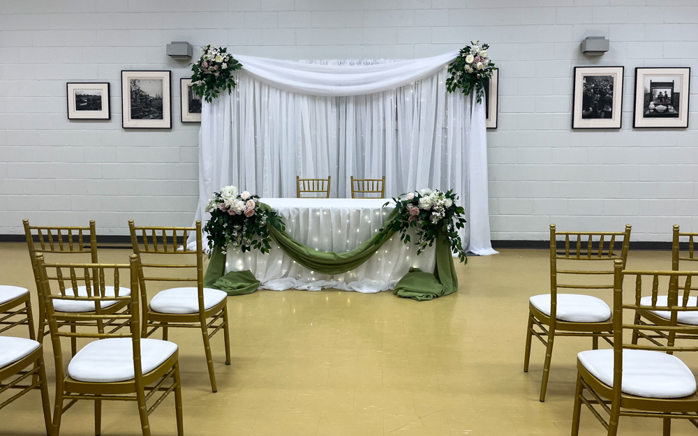 small head table with organza draping, mini lights, green accent fabric and flower bouquets. Organza backdrop with mini lights, chiavari chairs